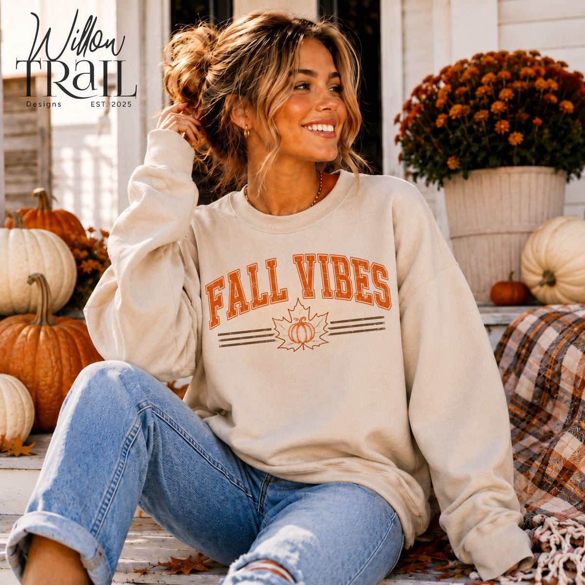 Model wearing a cream Fall Vibes crewneck sweatshirt with orange lettering and a pumpkin design, seated on a porch.