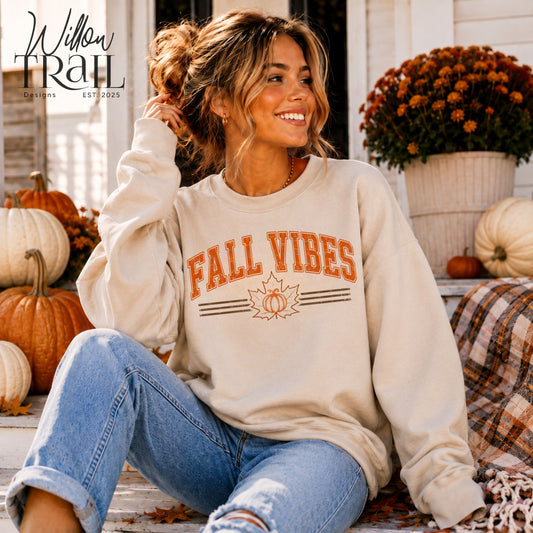 Model wearing a cream Fall Vibes crewneck sweatshirt with orange lettering and a pumpkin design, seated on a porch.