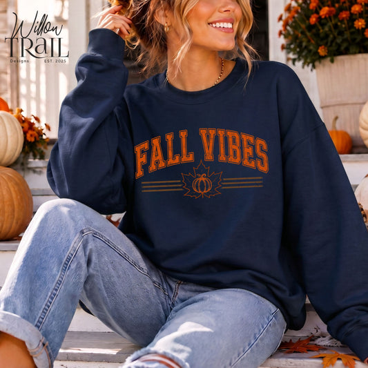Model wearing a navy Fall Vibes crewneck sweatshirt with orange lettering and a pumpkin design, seated on a porch.