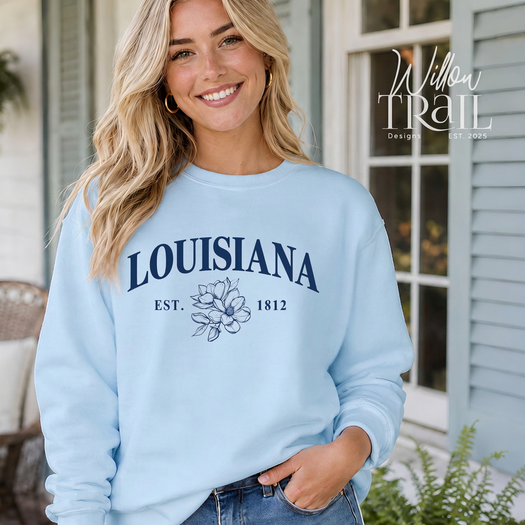 Model wearing a light blue Louisiana crewneck sweatshirt with navy lettering, a magnolia flower illustration, and EST. 1812 detail.